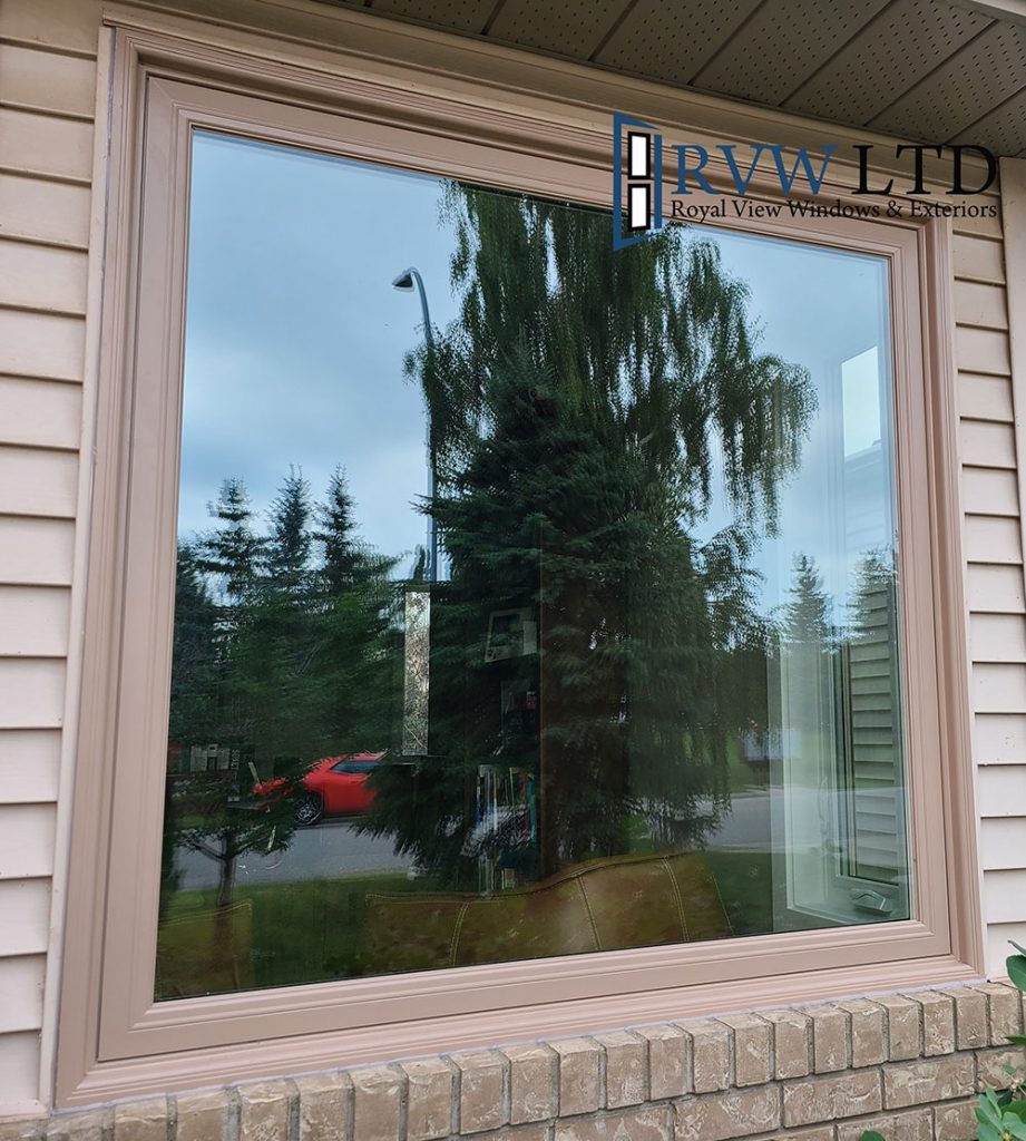 Large bay window with beige frame, reflecting trees and a red car. Siding and brick from the building are visible, along with the RVW LTD logo in the top right corner.