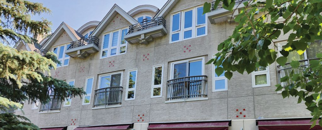 A beige, three-story townhouse with multiple windows and small balconies, partially obscured by green trees in the foreground.