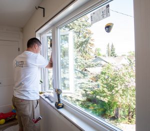 A man installs a new window in a residential home, using tools placed on the windowsill. Calgary Window & Door Manufacturers provide quality materials for the upgrade. Trees and houses are visible outside.