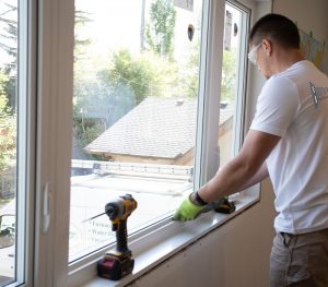 A person wearing safety glasses installs a window indoors using a power drill and screwdriver, with a Calgary Window & Door Manufacturers service van visible outside.