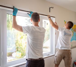 Two men from Calgary Window & Door Manufacturers install or adjust a large window in a bright room; one wears gloves while both work together to fit the frame.
