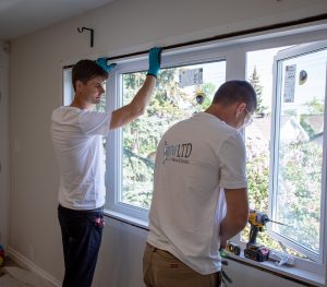 Two workers in white shirts install a window in a residential home; one holds the frame while the other uses a cordless drill, showcasing the expertise of Calgary Window & Door Manufacturers.