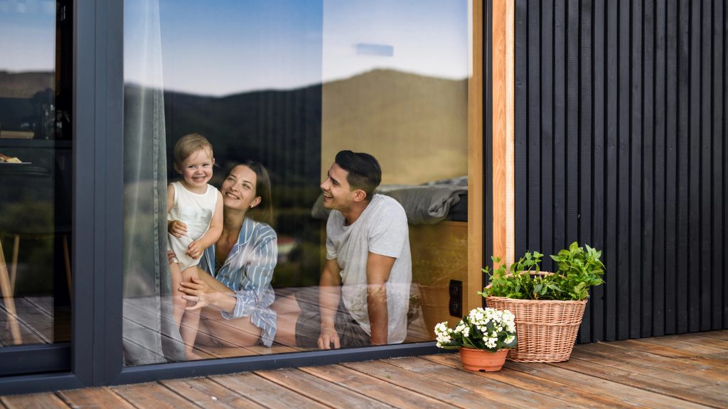 A smiling family with a toddler sits inside by a glass door, looking out onto a wooden deck with flowers and green plants—an ideal spot to learn how to spot leaky or damaged window frames in Calgary homes.