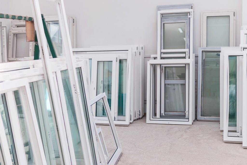Multiple white window frames and glass panels are stacked and leaning against the walls inside a bright room with a concrete floor. Are your doors hard to close? It might be time for a replacement.