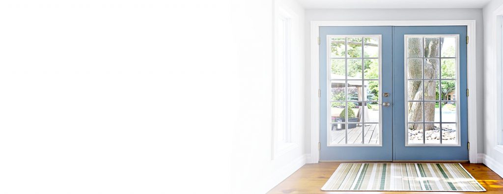 A pair of blue French doors with glass panes, expertly installed, open to a deck and trees outside; a striped rug lies on the wooden floor inside—showcasing quality Doors Replacement and Installation.