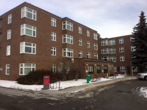 A four-story brick apartment building with rectangular windows, surrounded by sparse snow, bushes, and a few benches near the entrance.