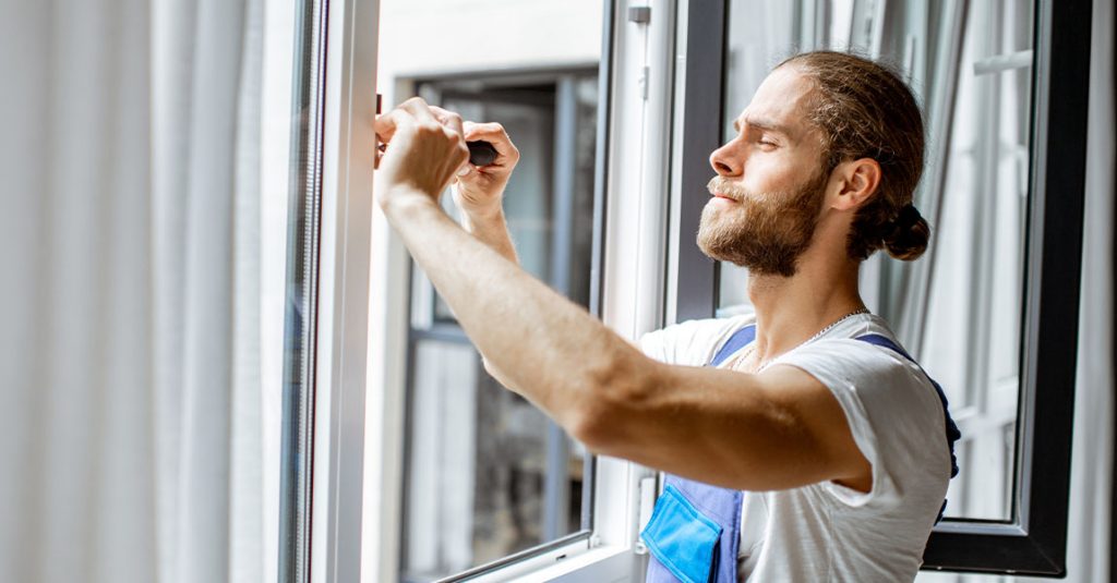 A man in work overalls uses a screwdriver to adjust or repair a window latch inside a bright room, showing how window installation affects energy bills in Calgary homes by ensuring proper sealing and efficiency.