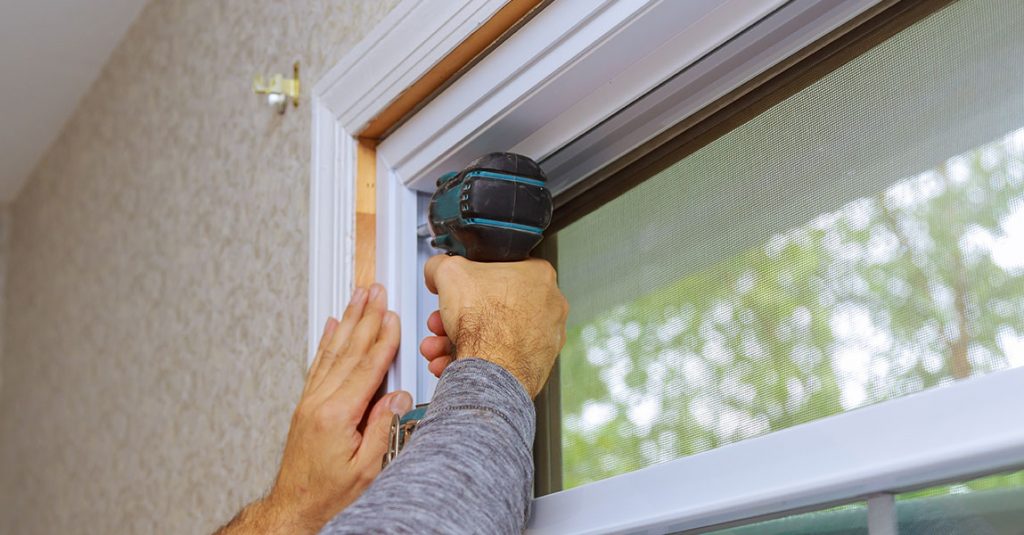 A person uses a power drill to install a window frame inside a house, demonstrating how window installation affects energy bills in Calgary homes by ensuring proper sealing and insulation.