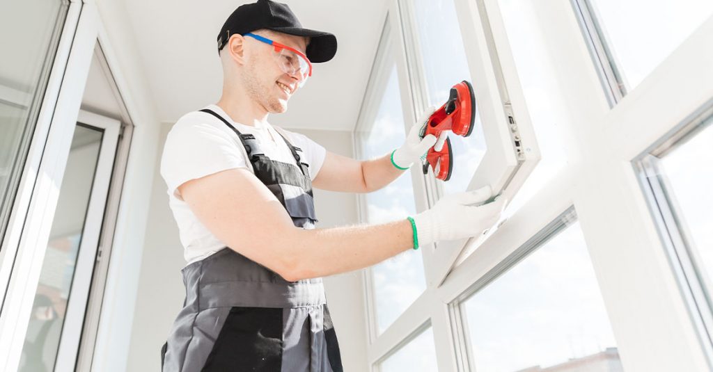 A worker in gloves and uniform installs a glass window using a suction tool in a bright room, demonstrating how window installation affects energy bills in Calgary homes.