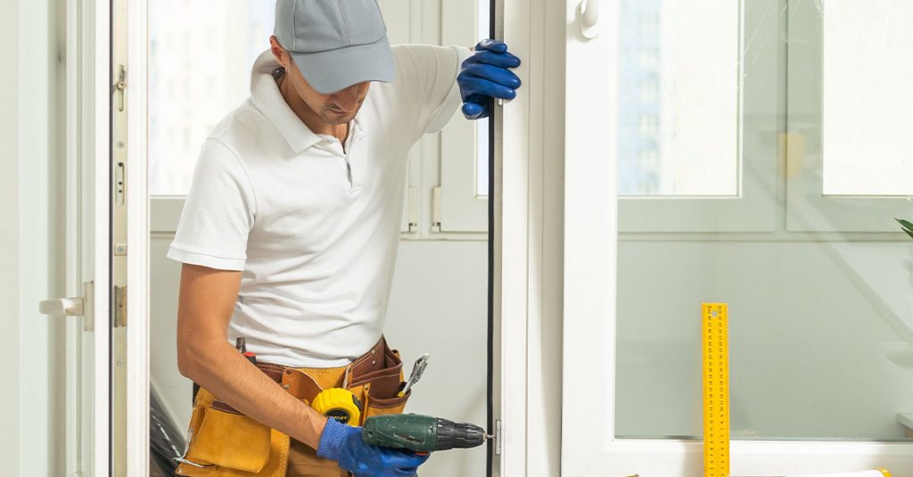 A worker wearing a cap, gloves, and tool belt uses a drill to install or repair a door in a bright indoor setting, highlighting how window installation affects energy bills in Calgary homes by improving efficiency.