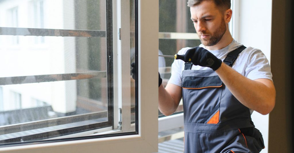 A man in work overalls and gloves installs or repairs a window using a screwdriver inside a building, highlighting how window installation affects energy bills in Calgary homes.