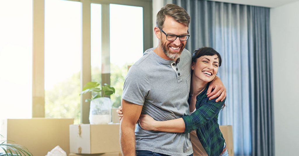 A smiling man and woman embrace in a bright room with unpacked moving boxes, a potted plant, and the peace of mind that comes from Why Our Window Replacements Include Full Frame and Insulation Checks.
