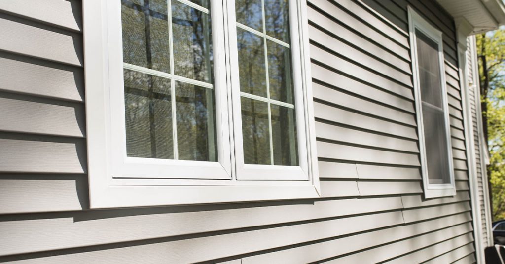 Close-up of a double window with white frames on the exterior of a house with light-colored vinyl siding. Trees are reflected in the glass, highlighting why our window replacements include full frame and insulation checks for lasting quality.