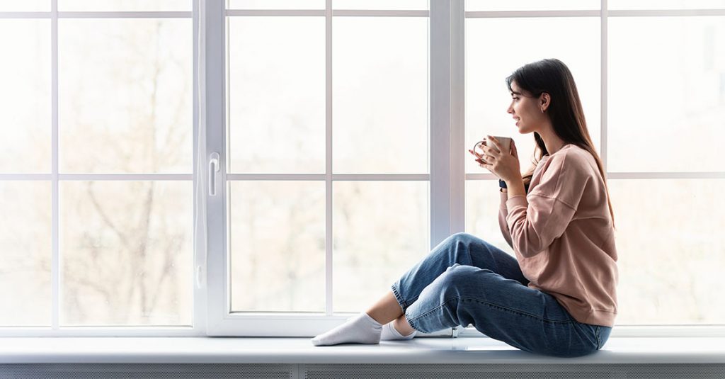 A woman sits on a windowsill, holding a mug and enjoying the view outside. With new windows that feature full frame and insulation checks, she relaxes in comfort, wearing a pink sweater, blue jeans, and white socks.
