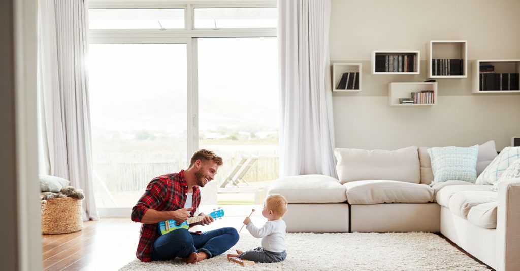 An adult and a baby sit on a living room floor; the adult holds a small guitar while the baby taps drumsticks. A white sofa and shelves form the backdrop, highlighting why our window replacements include full frame and insulation checks for cozy family moments.