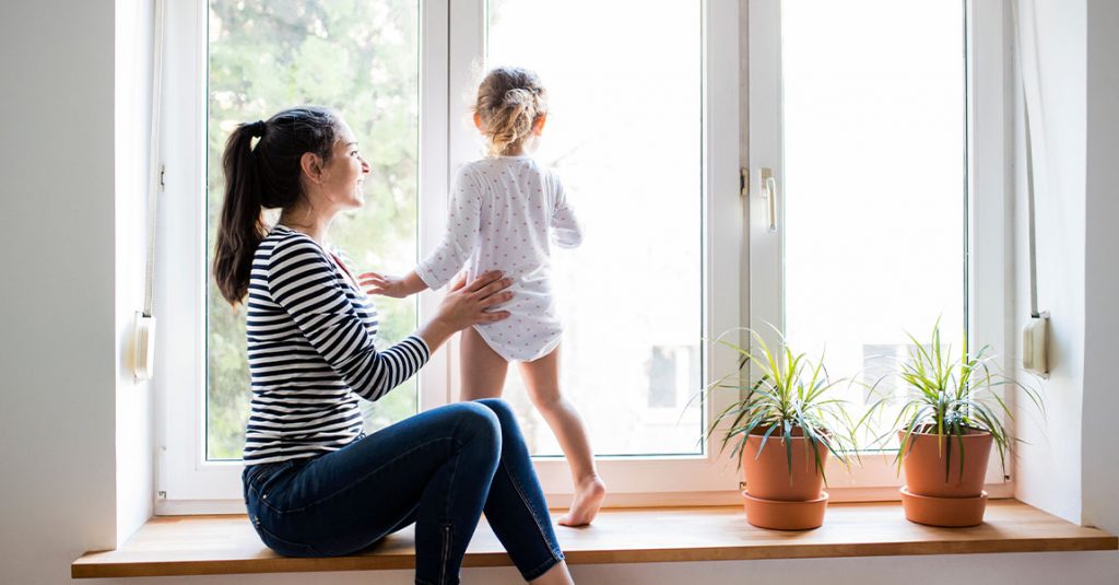 A woman supports a young child standing on a wide windowsill next to two potted plants, both looking outside through a large window—showcasing why our window replacements include full frame and insulation checks for comfort and safety.
