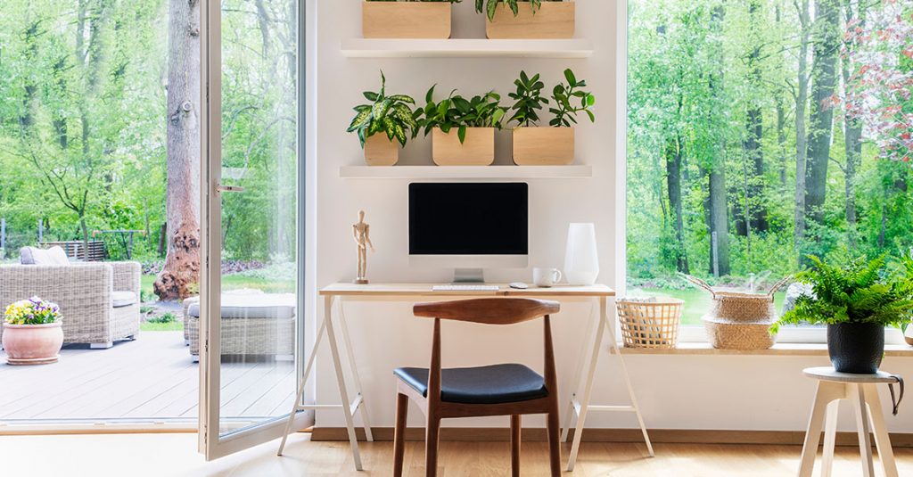 A modern home office with a desk, chair, computer, and potted plants on shelves sits by a large window—demonstrating why our window replacements include full frame and insulation checks for comfort and energy efficiency.