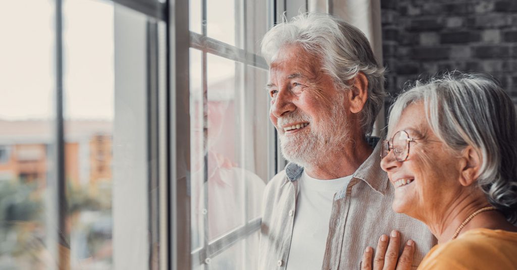 An older man and woman stand side by side, smiling as they look out of a window with natural light shining on them—showcasing why our window replacements include full frame and insulation checks for lasting comfort.