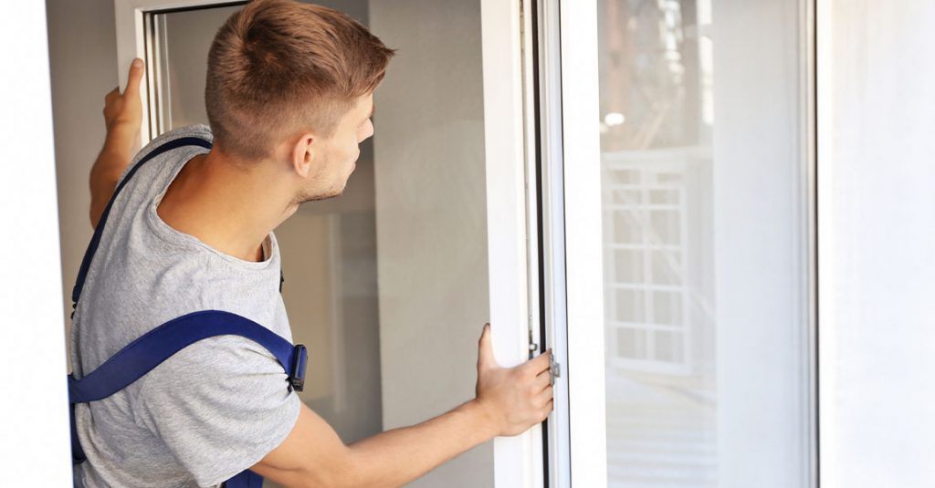 Person installing a white-framed vinyl window, wearing work overalls and a grey shirt, seen from the side inside a building—demonstrating why our window replacements include full frame and insulation checks for optimal performance.